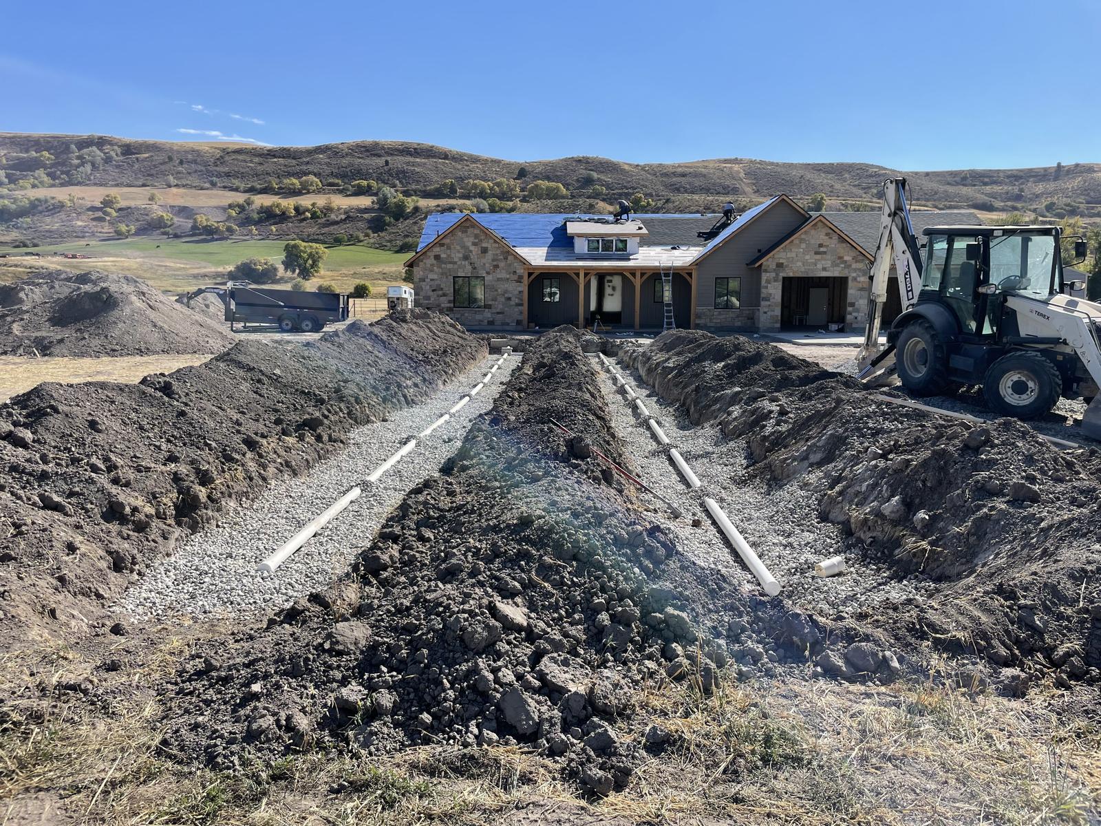 Open drain-field trenches lined with gravel and white distribution pipe, with a new stone-front home in the background
