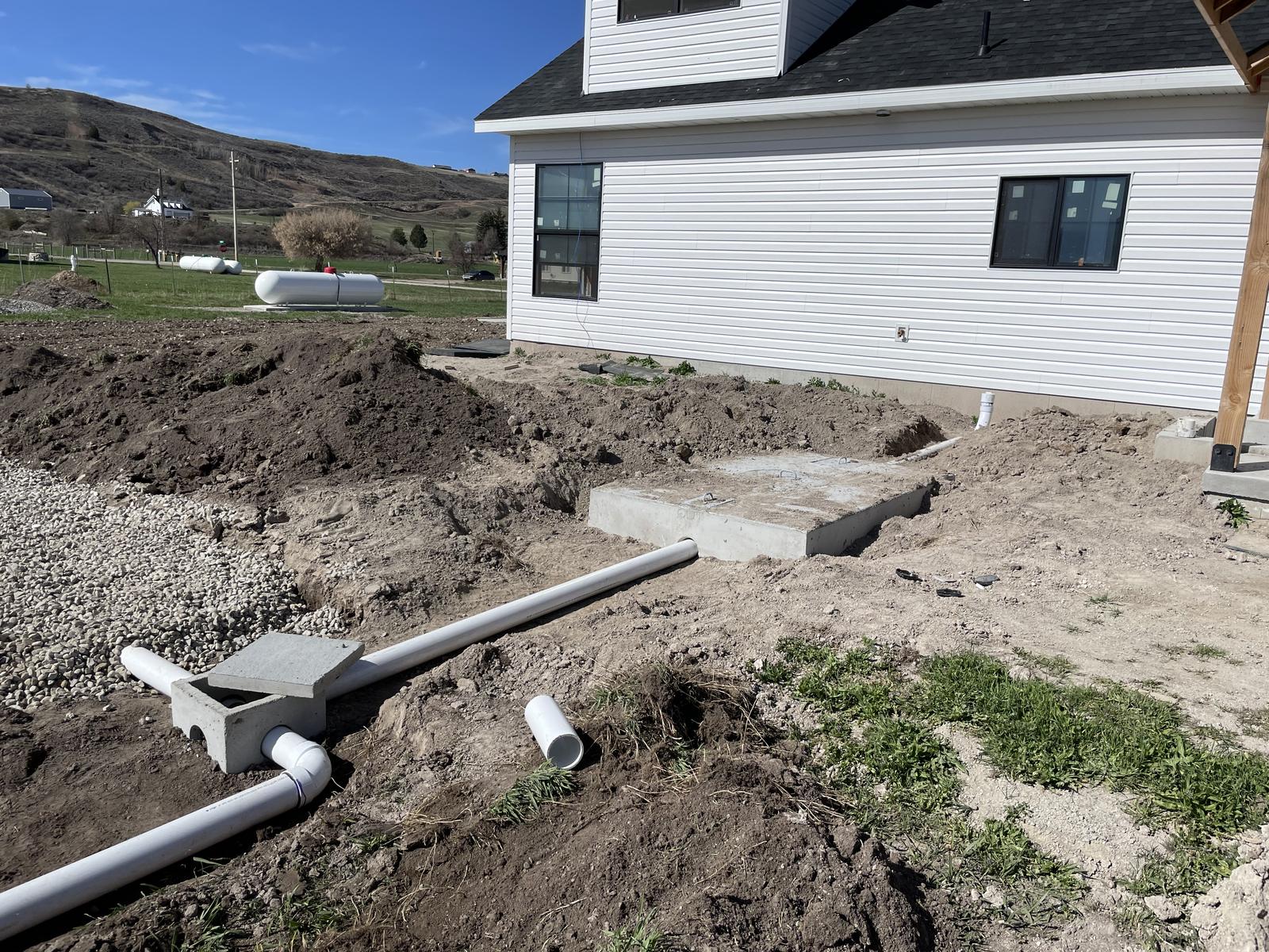 Septic tank lids beside a new home, with white PVC outlet line running to a small concrete box